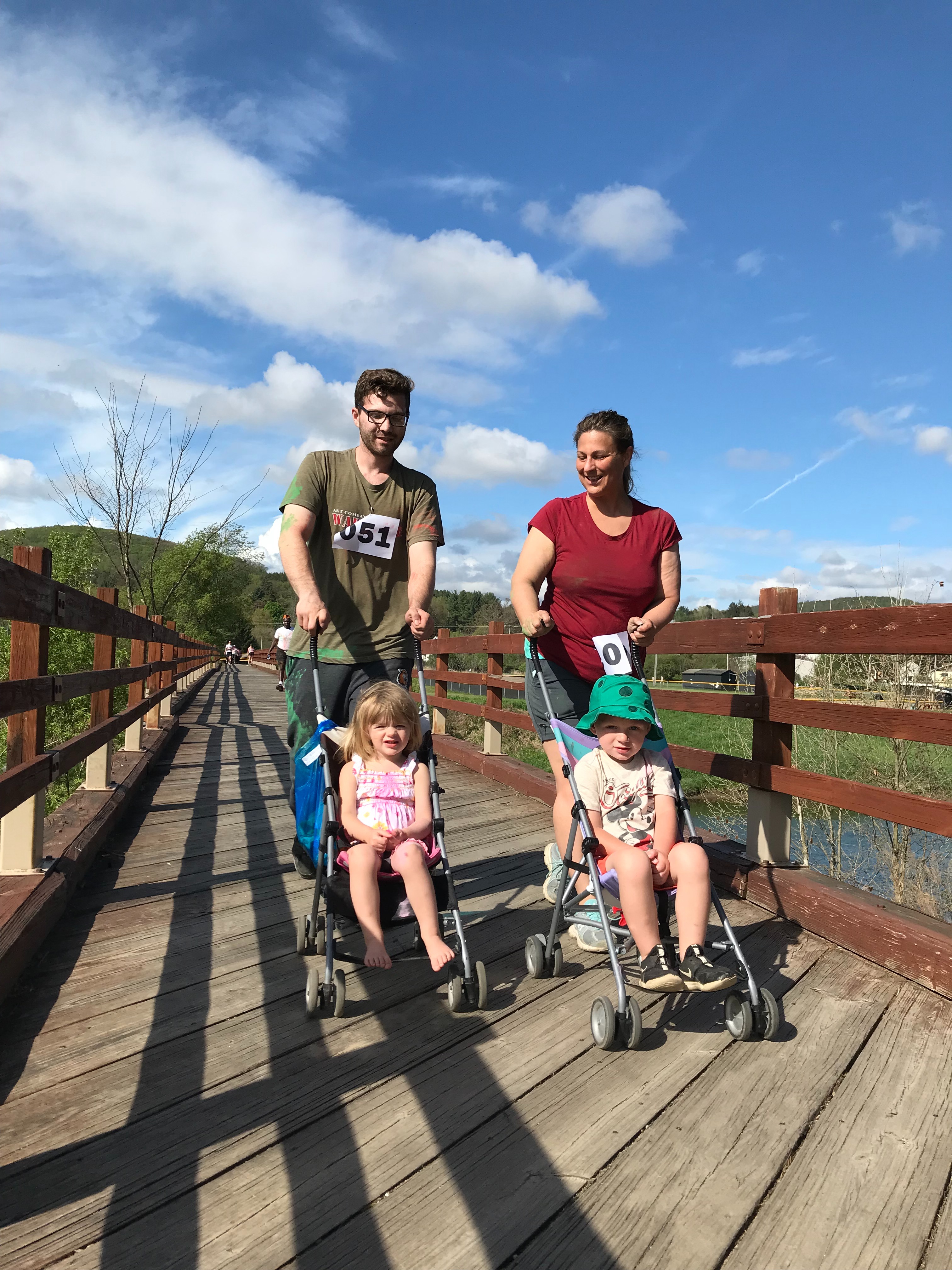 Man and woman pushing strollers with young children across a bridge during the Color Run/Family Walk